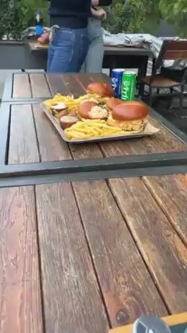The video shows a food photography shoot taking place outdoors. The focus is on a wooden table on which a metal tray of burgers and fries sits, along with small containers of sauce. In the background, other wooden tables are visible, one with beverages. Two people stand near the tables. A man in a teal sweater and blue jeans is preparing to film the food using a RONIN camera mounted on a stabilizer. A black coat with fur trim rests on a chair next to him. There are light stands visible in the background. The scene appears to be on a patio or in an outdoor dining area.