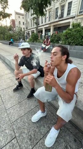 Two young men sit on a low stone wall in a public space. Both are smoking cigarettes. One wears a white tank top, light-colored shorts, and white sneakers. He turns his head to look directly at the camera and holds up his cigarette. The other man wears a baseball cap, a black T-shirt with white designs, light-colored shorts, and black sneakers. In the background, a Black man sits cross-legged on a similar stone wall and plays a drum. Other people sit and stand nearby in the background. Buildings with multiple stories line the street behind them. The sky is bright.