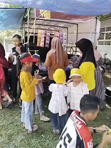 The video shows an outdoor gathering, likely a market or event, with a makeshift stall set up under a blue and white tarp. Several people, including children and adults, are present. In the foreground, two young girls are standing. One, wearing a yellow shirt and a red visor, appears to be holding something to her mouth. The other, in a white dress and yellow hat, is looking forward. Behind them, several other children and adults are visible, some interacting with the stall which displays various items. To the left, a group of people are gathered around a table, with some seated. The ground is covered in grass, and trees are visible in the background, suggesting a park or open outdoor area. The lighting indicates it is daytime.