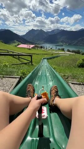The video is a first-person perspective of someone riding a toboggan down a green metal track that winds through a grassy hillside. The rider's legs and feet, clad in brown hiking boots, are visible in the foreground, and they are holding onto a black handle. The toboggan has a orange item attached to it. The track curves to the left, revealing a scenic view of a lake surrounded by mountains under a partly cloudy blue sky. In the distance, there are some buildings with pink roofs and a fence. The overall impression is a thrilling outdoor activity in a picturesque mountain landscape.