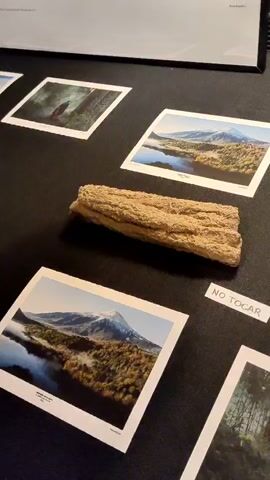 The video shows a close-up, high-angle, shaky shot of various items on a dark surface. Several printed photographs are visible, depicting landscapes with mountains and a person in a forest. There are also several irregular-shaped, light-brown objects, possibly rocks or pieces of wood, placed between the photographs. A small white label with the text "NO TOCAR" (Do Not Touch) in black letters is placed on the surface. In the background, a white lamp base is partially visible on the right. The video pans across the items from left to right.