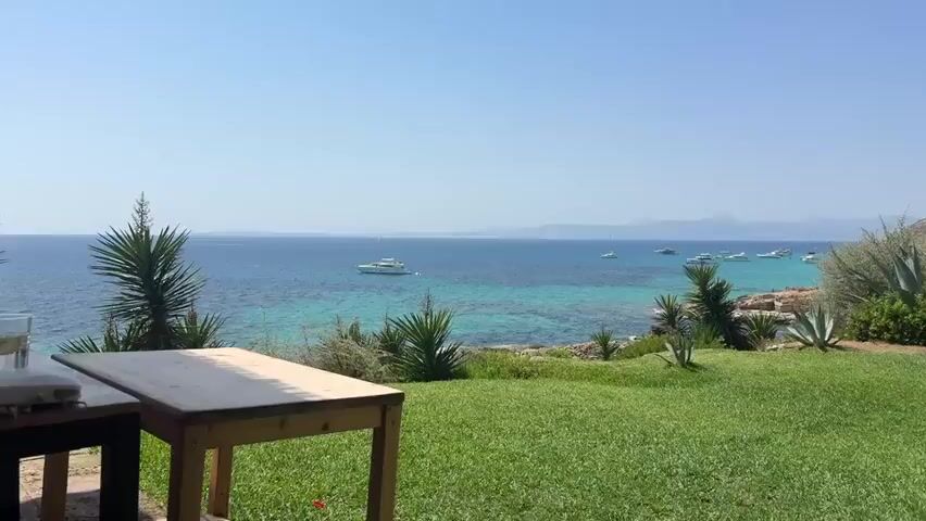 The video opens with a wide shot of a beautiful turquoise sea with several yachts anchored offshore, bordered by a green grassy lawn and lush vegetation. The camera then pans to the right, showing a manicured lawn with lounge chairs and umbrellas, and a few people relaxing. Next, the camera focuses on a table set for a meal outdoors, with wine glasses, water bottles, and menus. Two people, a man and a woman, are seated at the table. The man is wearing a white tank top with red trim, and the woman is wearing a light-colored dress. They appear to be engaged in conversation. The video continues with shots of the outdoor dining area, the surrounding greenery, and more people enjoying the scenery. The man smiles at the camera, and the woman gestures with her hand. The overall atmosphere is relaxed and leisurely.