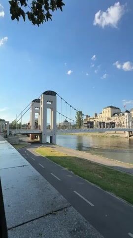 The video pans across a scene featuring a bridge, a river, and buildings. On the left side, a suspension bridge with white towers and cables is visible. A paved path, presumably for cycling or walking, runs alongside the river. In the middle ground, the river flows, with buildings lining the opposite bank. These buildings appear to be a mix of modern and classical architecture, with some featuring domes and columns. On the far bank, a few people are visible near the water's edge, one of whom is standing next to a bicycle. The sky is a clear blue with a few scattered clouds, and the sun appears to be shining, casting light on the scene. Further along the river, another bridge with multiple arches is prominent. On the near bank, a statue of a reclining figure is situated next to the paved path. The overall environment suggests a scenic urban waterfront setting.