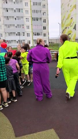 This video captures a group of children engaged in an outdoor activity, likely a game or a party, in a residential area. The subjects are primarily children of various ages, some adults are also visible. They are gathered around a large, colorful parachute that is being manipulated in a way that suggests a game is in progress. Some children are holding onto the edges of the parachute, while others are looking up, possibly at a ball or another object being thrown into the air. The setting appears to be a playground or a courtyard, with a tall, multi-story apartment building dominating the background. The building's architecture suggests a modern residential complex.

The activity appears to be energetic and fun, with many of the children showing expressions of excitement and engagement. The time of day seems to be daytime, and the weather appears to be overcast or slightly cloudy, as indicated by the lighting. The location is confirmed to be Фролы, Russia, which is consistent with the residential setting.

Notable details include the diverse clothing of the children, ranging from t-shirts and shorts to patterned skirts. One adult is wearing a bright purple jumpsuit, adding a touch of vibrancy to the scene. The presence of a red ball being tossed in the air is a key element of the game. The apartment building in the background has a somewhat uniform, repetitive design, typical of many housing developments. There is a playground structure visible in the distance, further reinforcing the idea of a community space for children. No visible text is present in the video.