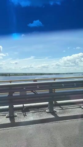 A view from a vehicle traveling over a bridge. The sky is blue with scattered white clouds. A wide body of water stretches across the horizon, with land and some trees visible in the distance. A metal railing with yellow accents runs along the side of the bridge. The road surface is asphalt with a yellow dividing line. The shadow of the railing is cast on the road. A light pole is visible on the bridge.