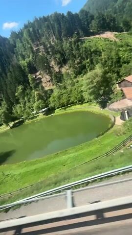 This media file captures a scenic view of a tranquil green lake nestled amidst lush, tree-covered mountains under a bright, sunny sky. The perspective suggests the footage is taken from a moving vehicle, possibly a train, as indicated by the blurry railing in the foreground.

The primary subjects are the natural landscape: the vibrant green lake, the dense evergreen forest blanketing the mountainside, and the expansive blue sky with scattered white clouds. A lone, large tree stands prominently on the grassy bank of the lake, its branches casting a shadow on the water. Several small buildings, possibly cabins or sheds, are visible along the lake's edge, adding a touch of human presence to the otherwise natural setting. The overall scene evokes a sense of peace and natural beauty.

The time of day appears to be midday, given the bright sunlight and the distinct shadows. The weather is clear and pleasant, contributing to the idyllic atmosphere. Location cues point to a mountainous region, likely in Italy given the provided context of Karneid - Cornedo all'Isarco, known for its natural landscapes.

There is no discernible activity or event taking place beyond the passive observation of the landscape. No people are clearly visible or engaged in any action. The dominant emotion conveyed is one of serenity and appreciation for nature's beauty. No text is visible within the frame.