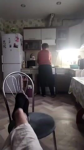 The video shows a person sitting in a chair in a kitchen with their feet propped up. The person is wearing socks and light-colored pants. In the background, another person, a woman wearing an orange shirt and a dark skirt, is standing at a kitchen counter, appearing to be cooking or cleaning. The kitchen is small and appears to be lived-in, with a refrigerator, cabinets, a microwave, and a table covered with a floral tablecloth. The lighting is dim, with a bright light coming from under the kitchen cabinets.