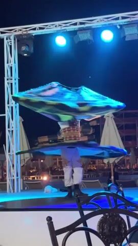 A man performs a whirling dervish dance on a stage at night. He is wearing white pants and a colorful vest, and two large, spinning skirts of differing colors. He spins rapidly while the skirts swirl around him, creating a colorful blur. The stage is illuminated with blue lights. Behind the stage, there are lounge chairs, umbrellas, and some sort of scaffolding. A few black chairs with ornate backs are visible in the foreground. The sky is dark.