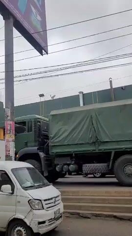 The video shows a military truck with a green tarp covering its cargo, driven by an unseen driver. The truck is moving on a street, passing by other vehicles such as a white van, a yellow taxi, a red and blue auto rickshaw, and a red car. The scene appears to be in an urban area with buildings and power lines visible in the background. The sky is overcast.