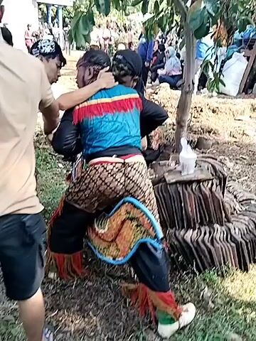 The video shows a group of people gathered around an individual in traditional Indonesian attire, potentially a performer. The person is wearing a colorful vest with fringe, patterned pants, and a head covering. Two individuals are close to the performer, appearing to adjust or apply something to their head or face.  Several other people are standing nearby, observing. The setting appears to be outdoors, possibly a village or rural area, with trees and other people in the background. There's a small table or platform nearby covered with tiles or plates. The atmosphere seems communal, possibly related to a cultural event or preparation for a performance.