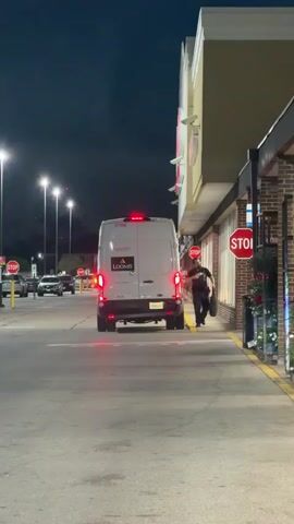 At night, a white Loomis van is parked in front of a building with a red logo. A man in a dark uniform and cap is walking towards the building with a bag. A woman in jeans and a gray shirt walks past the van in the opposite direction. The area is well-lit by streetlights, and there are several stop signs visible. Other cars are parked in the background. The building appears to be a retail store or possibly a bank based on the security presence.