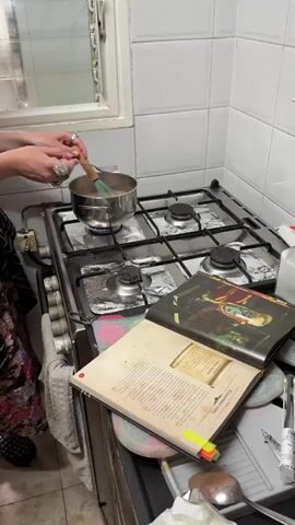 Here is a concise description of the video:

A person is stirring a liquid in a small pot on a gas stovetop in a kitchen. The stovetop is covered with foil. A large cookbook is open on the counter beside the stove, with several sticky notes marking pages.  The kitchen has white tiled walls and a window above the stovetop. The person is wearing a floral dress and rings. The scene is brightly lit.
