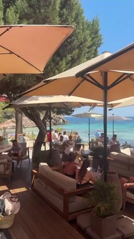 The video shows a crowded outdoor cafe with a beach and ocean in the background. Large beige umbrellas provide shade for the tables and seating areas. Several people are seated at tables and on couches, some are eating and drinking, while others are talking. A server walks by carrying a tray of drinks. In the background, the ocean is visible with people swimming and boats on the water. The overall scene depicts a lively and sunny day at a beachside establishment.