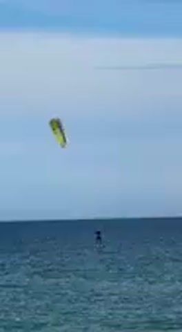 The video captures a wide shot of a person engaging in a water sport on a bright, sunny day. A individual is seen standing on a board in the deep blue ocean, propelled by a vibrant yellow and green kite flying high in the clear, light blue sky. The sky features a few subtle, wispy clouds, and the horizon line is visible in the distance. The water's surface shows gentle ripples, indicating calm conditions suitable for kitesurfing. The scene depicts an active, outdoor activity under favorable weather conditions. No text is visible in the frame.
