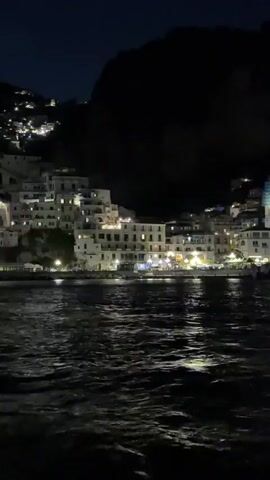 The video shows a night view of a town by the water. The buildings are mostly white and illuminated with lights, clustered together on a hillside. Dark water ripples in the foreground, reflecting the town's lights. A dark mountain rises in the background, obscuring the sky. The camera appears to be on a boat moving slowly through the water, with the view slightly shaking.