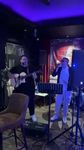 Two men are performing in a dimly lit room. The man on the left is playing an acoustic guitar and singing. He is wearing a black t-shirt and pants, and white sneakers. The man on the right is singing into a microphone. He is wearing a white t-shirt and pants, a black jacket, and white sneakers. There are two speakers on stands behind them, as well as music stands and a sound mixer. The room has black walls and a red curtain backdrop. There is a chair in the foreground and a ceiling fan overhead. The lighting is primarily blue.