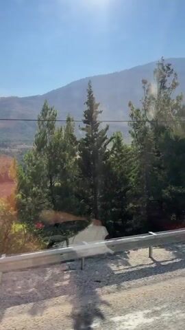 The video shows a view from a moving vehicle on a sunny day. The foreground consists of a road with gravel and some shadows, bordered by a metal guardrail. Beyond the guardrail, there is dense green foliage, including trees and bushes. In the background, a large, hazy mountain dominates the landscape, with a clear blue sky above. The sun is visible, casting a bright glare in the upper part of the frame. Electrical poles and wires can also be seen in the mid-ground. There are no people visible in the video.