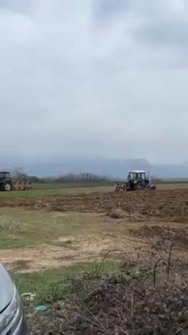 Two tractors are plowing a field on an overcast day. The field is largely brown dirt, with patches of green grass.  In the distance, trees and mountains are visible beneath a gray sky.  The tractors are some distance apart. The tractor on the left is primarily red; the tractor on the right is mostly light blue.  The foreground shows the edge of a silver or light-colored vehicle and some shrubbery.