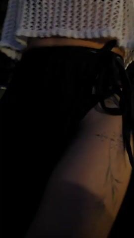 The video shows a person dancing outside at night. They are wearing a white, loose-knit sweater, a black skirt with a side tie, and appear to have a tattoo on their leg. The background is dark with some foliage visible. The person raises their arms and smiles as they move to music.