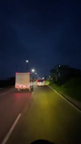 The video shows a first-person view of a vehicle driving on a road at night. The sky is dark blue, and streetlights illuminate the road ahead. A white truck with red taillights is driving in front of the viewer's vehicle. The road has a painted white line on the left side and a solid yellow line on the right, indicating traffic flow. Buildings are visible on the right side of the road, and trees and bushes line the left side. The viewer's vehicle casts a yellow light on the road directly in front of it.