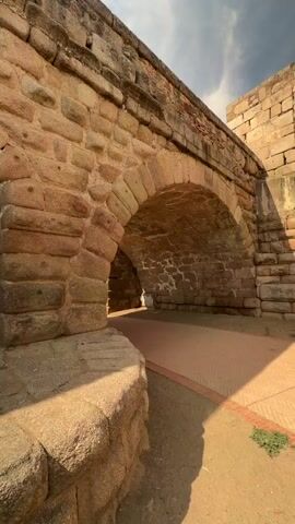 The video shows a low-angle, close-up view of an ancient stone bridge with multiple arches. The bridge is constructed from large, rough-hewn stones. Sunlight streams from the top right, casting shadows and highlighting the texture of the stones. The camera pans slowly to the left, revealing the interior of one of the arches, which appears to be a passage. Further to the left, another arch reveals a body of water, likely a river, reflecting the sky and greenery on the opposite bank. The path beneath the arches is paved with reddish-brown tiles. The overall impression is one of historical significance and weathered resilience.