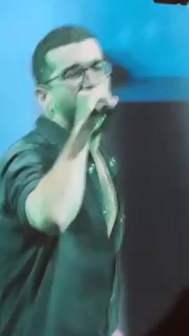 A man with a beard and glasses is singing into a microphone on stage. He is wearing a black shirt that is open at the collar. The lighting on stage is green.