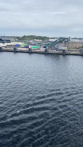 The video shows a wide shot of a port with several cargo containers on the dock. There are also some trucks and a large metal structure with a ramp. The water in the foreground is dark and choppy, with some small waves. The sky is cloudy. The video appears to be taken from a moving vessel.