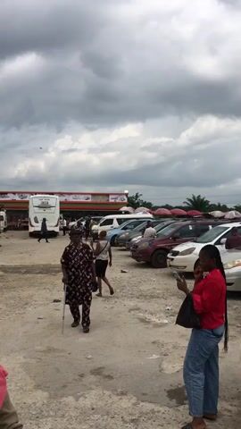 The video shows a busy outdoor scene, likely a transportation hub or market. In the foreground, two women are visible. The woman on the left is wearing a purple headscarf and a pink garment draped over her shoulder. The woman on the right is elderly and walks with a cane, wearing a patterned dress and headscarf. Several vehicles are present, including white minivans, a large white bus with tinted windows, and other cars. The ground is unpaved and appears to be dirt and gravel. In the background, a building with a red and green awning is visible, possibly a shop or service station. Some people are seen walking around and interacting. The sky is overcast with heavy clouds. The overall impression is of a bustling, possibly commercial, area in an outdoor setting.