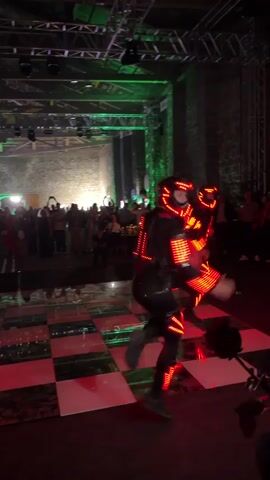 This video shows a performance by dancers in illuminated costumes, likely at a formal event or club in Portorož, Slovenia. The performers wear full-body suits that are outlined and patterned with bright LED lights in various colors, including red, blue, green, and purple. The lights change color and pattern throughout the performance, creating a dynamic visual effect.

The dancers are on a checkered dance floor, which also appears to be illuminated. In the background, a crowd of people can be seen watching the performance, some with their hands raised. The setting seems to be a large, industrial-looking indoor space with exposed brick walls and exposed ceiling trusses with lighting equipment. The lighting is dramatic, with green uplighting on the walls and spotlights directed at the dance floor.

The dancers are performing energetic moves, suggesting a choreographed routine. The overall mood of the video is festive and exciting, with the illuminated costumes and upbeat music creating an engaging atmosphere. The time of day is not explicitly stated, but the indoor setting and artificial lighting suggest it is likely evening or night. There is no visible text in the video.