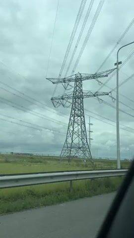 A drone flies low over a highway, capturing a wide shot of a massive metal power tower against a cloudy sky. The drone then pans right, revealing a street lamp on the right side of the frame. As the drone continues, it passes under a concrete bridge with graffiti on its underside. The bridge's ceiling is adorned with large purple letters and other graffiti tags. The drone continues to move forward, providing a view of the graffiti-covered bridge from beneath.