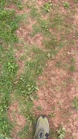 The video shows a person walking on dry dirt and grass towards a CF MOTO ATV. The ATV is parked on a dirt path with green grassy fields and hills in the background. The ATV is black and gray camouflage.
