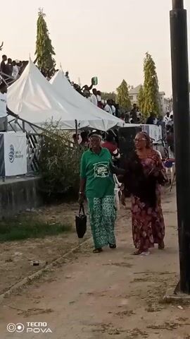 The video shows an outdoor event taking place in a dusty area with sparse vegetation. There are crowds of people, white tents, and a grandstand filled with spectators. Two women are walking along a path, one wearing a green shirt and patterned pants, and the other wearing a long, patterned dress with a dark shawl. The grandstand is decorated with banners and logos, and there are people standing and waving on top of it. A group of people in yellow vests are sitting on chairs next to the edge of the playing field. Backpacks and other items are strewn on the ground near them. The video is shot from a ground-level perspective with the camera moving slightly. The sky is light and the atmosphere appears to be warm.