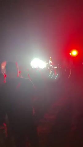The video displays a crowd of people at an outdoor event at night, illuminated by stage lights and red flashing lights. In the foreground, a person in silhouette faces away from the camera, holding a can in their left hand. Green lights are visible on their lower legs. The middle ground shows a dense crowd of people facing a brightly lit stage with performers. The background consists of the dark night sky and more lighting equipment. The scene appears to be a concert or festival.