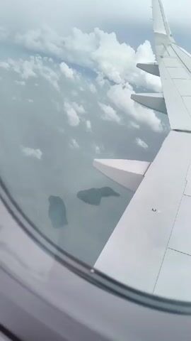An aerial view from an airplane window shows the wing of the plane and islands in the sea below. The sky is cloudy and the sea is calm.