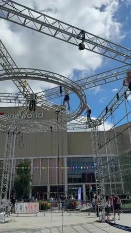 The video shows an outdoor high ropes course. A person in athletic wear is navigating the course while attached to safety lines. The course is constructed from metal scaffolding and suspended ropes and elements. In the background, there is a Cineworld building and a Starbucks. There are also people on the ground watching the person on the ropes course. The sky is blue with some clouds.
