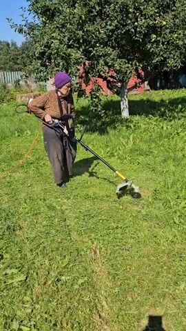 An elderly woman wearing a purple headscarf, a patterned brown long-sleeved shirt, and dark pants is using a string trimmer to cut grass in a yard. She is holding the trimmer with both hands and appears to be moving it back and forth. The trimmer is connected to an orange electrical cord that runs to the left of the frame. The yard is covered in green grass, some of which has been recently cut. Behind the woman, there is a tree with green leaves and a red building with a wooden fence. The sun is shining, casting shadows on the ground.