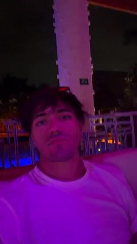 A person with dark hair, wearing a white t-shirt, is seen drinking from a clear glass with a straw in a dimly lit outdoor setting. The person appears to be sitting or reclining on a cushioned area with a red backing. The lighting is primarily purple, with blue and white accents. In the background, there are outdoor tables and chairs. A tall, white, textured structure is also visible in the background. The camera angle shifts slightly during the video.