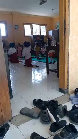 A group of children, mostly young girls, are kneeling or sitting on a green mat on a tiled floor, presumably in a classroom. They are all wearing school uniforms, which consist of white shirts and maroon skirts or pants. Several of the girls are wearing hijabs. The children appear to be engaged in some sort of activity, possibly a dance or a group exercise, as they are all positioned in similar poses and some have their hands raised or in motion. The room has yellow walls and wooden trim. There are windows on one wall, and some furniture, including a desk and shelves, are visible in the background. A fan is positioned on the left side of the frame, and shoes are scattered on the floor in the foreground, suggesting an entrance or exit area. The lighting appears to be natural, coming from the windows.