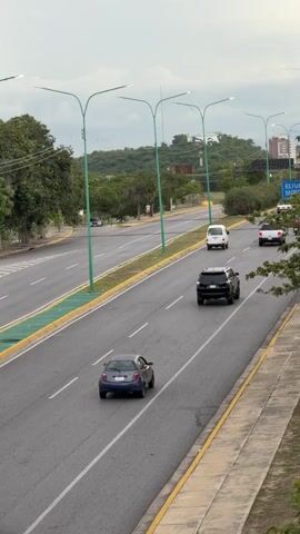 A drone shot surveys a multi-lane asphalt road. The road has white lane markings, a yellow painted curb, and a green painted lane. The scene is populated with various vehicles including a white SUV on the left, a blue sedan and a black SUV in the middle, and a white van and a black truck on the right. There are also two motorcycles, one with a rider in a red jacket and the other with a rider in a blue jacket, moving along the road. Trees and lampposts line the sides of the road, and a billboard with the word "REFUGIO" is visible in the distance. The sky is overcast.