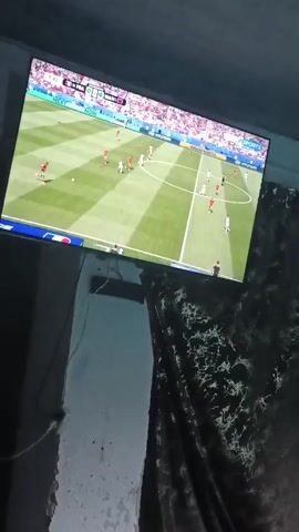 The video shows a television screen displaying a soccer match. The score on the screen reads 0-0 with 10 minutes into the match. A person is then shown playing pool. They are leaning over a green pool table, holding a pool cue, and about to strike a white ball. There are three pendant lights hanging from the ceiling above the pool table.