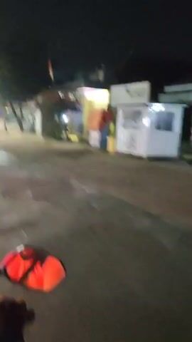 The video begins with a nighttime street scene. The camera pans from left to right, showing a blurry image of a road and sidewalks. There are lights from buildings and streetlights, casting shadows and reflections on the wet pavement. A red object, possibly a traffic cone, is visible on the left side of the frame.

As the camera continues to pan, a red car with its headlights on drives across the road from left to right. The car is moving quickly, and its taillights are visible as streaks of red. Behind the car, a building with a sign that reads "SIGNATURE" is partially visible.

The scene appears to be a commercial area with a few shops and businesses. The lighting suggests it is nighttime or late evening. The overall impression is of a transient, possibly busy, urban environment.