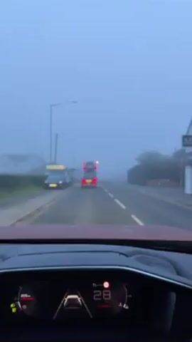 The camera is in a car, providing a driver's perspective while driving on a road shrouded in dense fog. The car's dashboard is visible, showing a speed of approximately 28 km/h. Ahead, a double-decker bus and a red car are driving in the same direction. The road markings are visible despite the fog, and there are buildings on either side of the road. Streetlights illuminate the scene with a faint glow. There is music playing in the background. The overall atmosphere is heavily obscured by fog, limiting visibility.