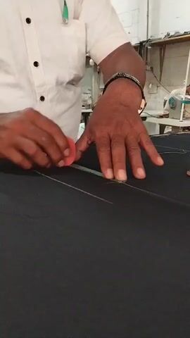 A person wearing a white collared shirt and a dark bracelet on their left wrist is seen from the chest down, standing at a table and marking a large piece of dark fabric. They hold a small red piece of tailor's chalk in their right hand and use a long, narrow green ruler as a guide to draw a straight line. After drawing one line, they pick up a green piece of chalk and use the same ruler to draw another line parallel to the first. In the background, another individual is seated and operating a sewing machine. The environment appears to be a workshop with multiple sewing machines visible.