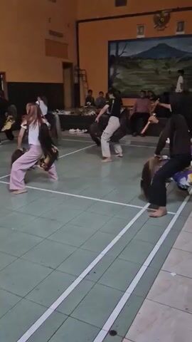 A group of people, some wearing black tops and pants or skirts, are performing a dance inside a room with yellow walls and a mural of a mountain scene. The floor is green with white lines, resembling a basketball court. Several performers are crouched down, wearing what appear to be horse costumes made of fabric with long, flowing manes. Some individuals are holding sticks. In the background, there are spectators sitting and standing, some appearing to be musicians with drums and other instruments. The lighting is dim, with some overhead lights visible.