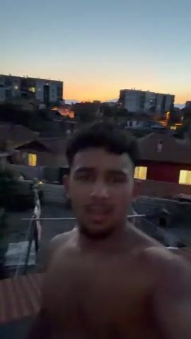 A young man with curly dark hair and a bare chest is recording a video of himself against a backdrop of a sunset over a residential area. He is smiling broadly, with his mouth open in an excited expression. The buildings in the background are blurred due to the camera's movement. The sky displays a gradient of orange, pink, and blue, indicating the sun is setting.