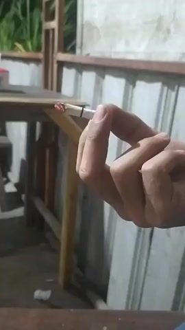 A person's hand holds a lit cigarette between their thumb and forefinger. The cigarette is in focus, with its glowing end visible. In the background, a wooden table and a corrugated metal wall are out of focus. The lighting suggests an outdoor setting at dusk or in a dimly lit area.