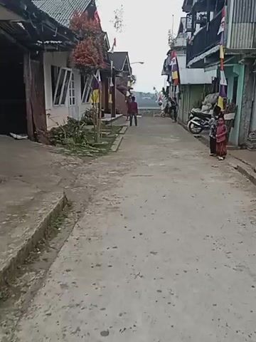 A person on a scooter travels down a narrow street past houses and other people. The street is made of concrete and is lined with buildings on either side. There are flags hanging from the buildings, and a motorcycle is parked on the right side of the street. Several children are standing on the right side of the street. One person is kicking a ball.