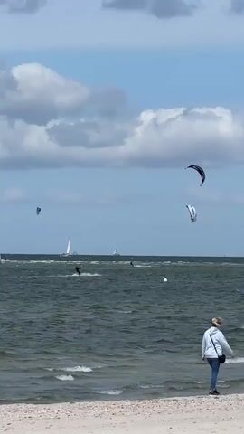 A video displays a beach with calm waves lapping against the shore. In the distance, several people are kiteboarding on the water under a bright blue sky with fluffy white clouds. There are also a few sailboats visible. A woman is walking along the beach in the foreground.