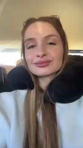 A woman with long brown hair and sunglasses on her head is shown from the chest up, wearing a white sweatshirt and a black neck pillow.  She is smiling slightly at the camera. A glimpse of the interior of a car is visible behind her. Music is playing.