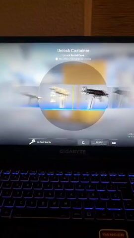 The video shows a close-up of a laptop screen displaying the Counter-Strike: Global Offensive (CS:GO) game interface. The screen shows the "Unlock Container" menu for the Recoil Case. The roulette-style selection is in progress, showing blurred images of various weapon skins. The spinning stops on a Negev | Drop Me skin. The background shows a plane and airport baggage. The laptop is a Gigabyte brand, with its RGB keyboard lit up in blue. A "DANGER" sticker is visible on the bottom right corner of the laptop. The video appears to be a recording of someone opening a CS:GO weapon case.